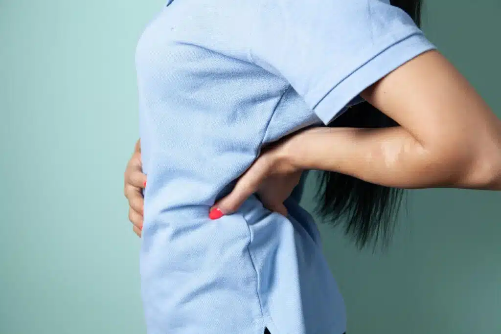 Person in blue shirt holding lower back with both hands against a teal background